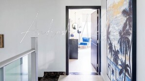 Um corredor moderno com guarda-corpo de vidro, uma grande pintura de palmeiras e um vislumbre de uma sala de estar com sofá azul.
