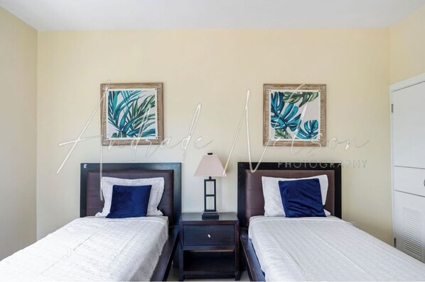 A hotel room with two beds, each with a headboard, a nightstand with a lamp, and framed tropical leaf artwork on the wall.