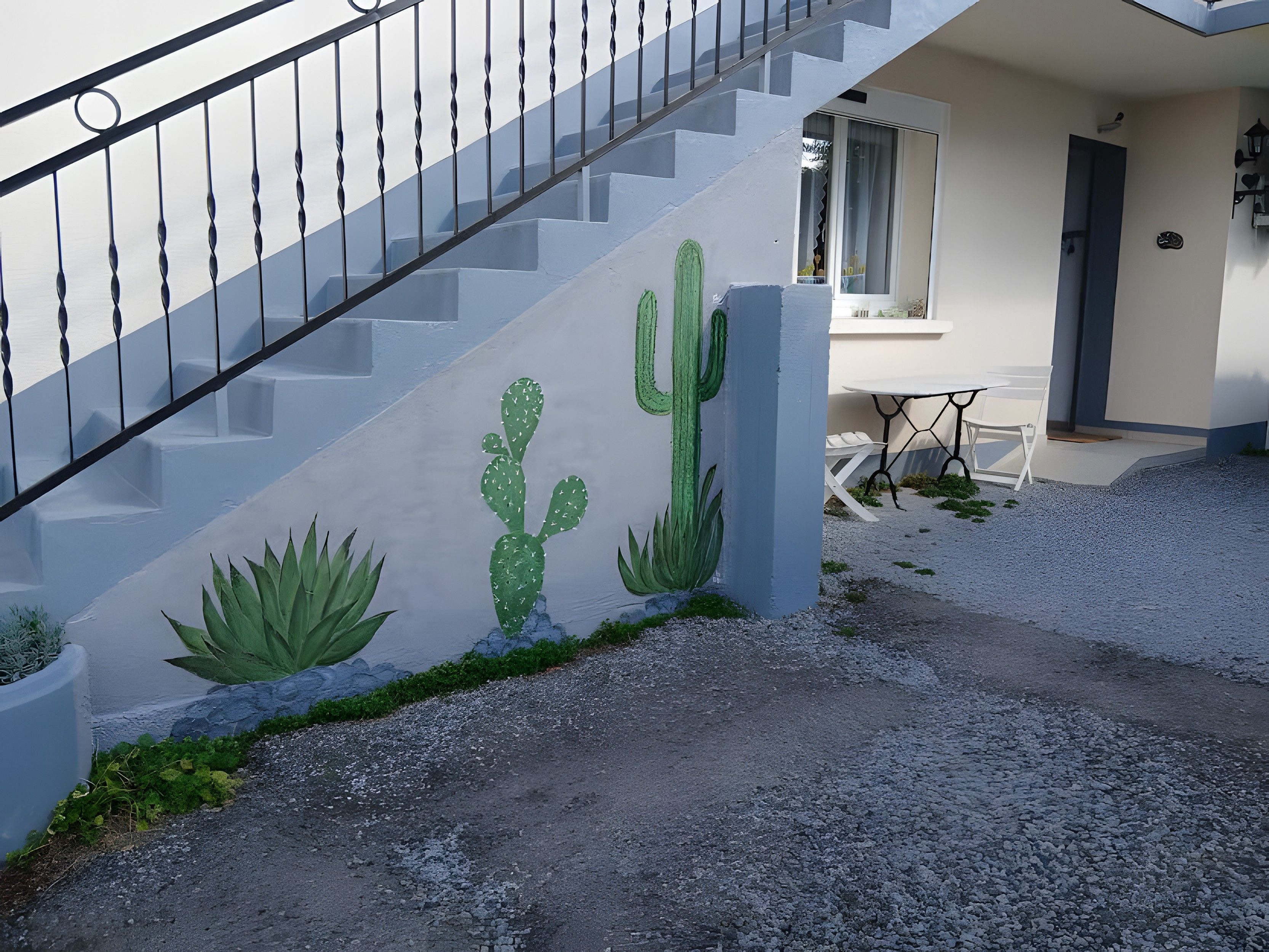 Una escalera con barandilla de metal, una pared con un mural de cactus y un pequeño espacio al aire libre con una mesa y sillas.
