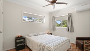 A bedroom with a bed, two windows, a ceiling fan, a nightstand, a lamp, a chair, and a small table. - Roderick Street 83, Moffat Beach (Moffat Beach)