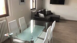 Un salon moderne avec une table à manger à plateau en verre, des chaises blanches, un canapé gris et un téléviseur à écran plat fixé au mur.
