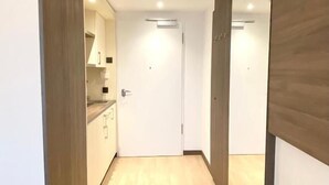 Quarto moderno e minimalista com piso de madeira, porta branca e área de cozinha visível através de uma porta aberta.