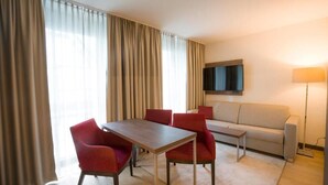 Quarto de hotel com mesa de jantar, quatro cadeiras vermelhas, sofá, televisão fixada na parede e abajur.