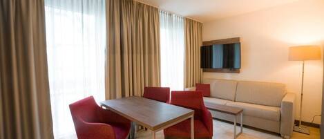 A hotel room with a dining table, four red chairs, a sofa, a TV mounted on the wall, and a floor lamp.