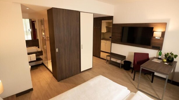 Quarto de hotel com cama, escrivaninha, cadeira, televisão e cozinha compacta.