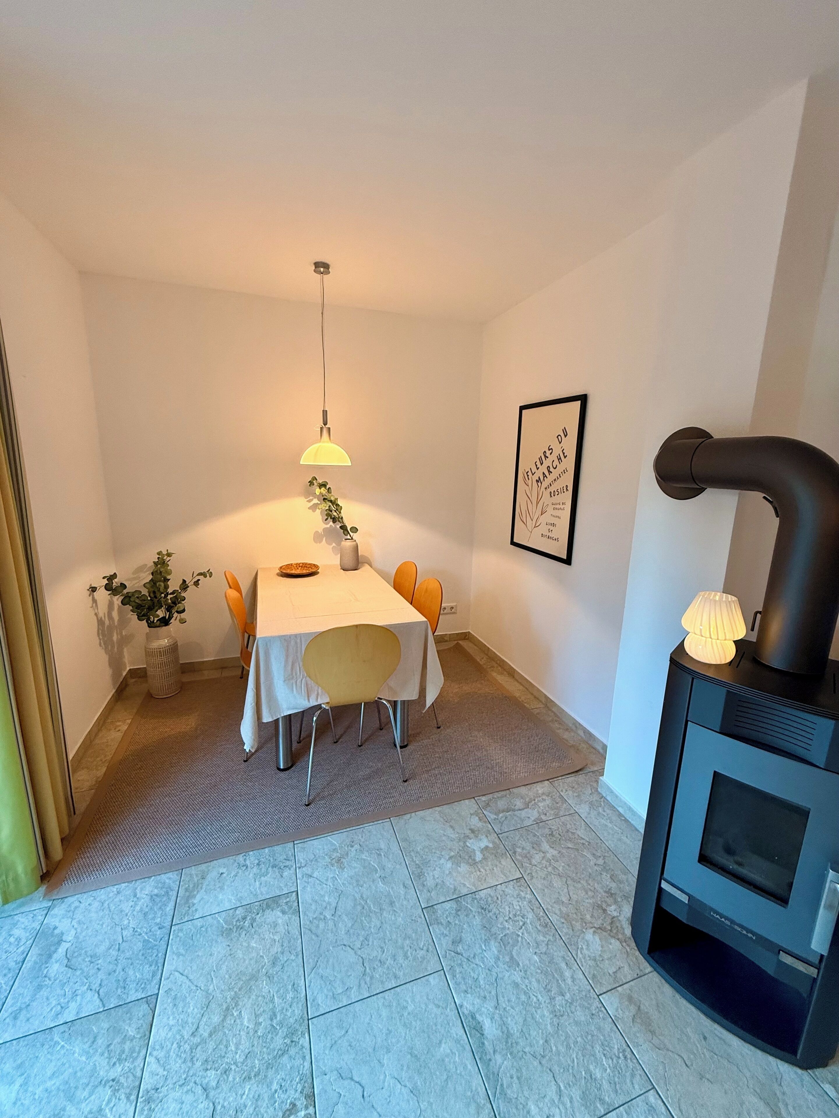 Ein Essbereich mit einem Tisch für vier Personen, einer Hängeleuchte, einem gerahmten Bild an der Wand und einem Kaminofen.