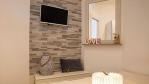 Un baño moderno con pared de azulejos, televisor montado, tocador con espejo y elementos decorativos, incluyendo un florero y un letrero que dice "LOVE".