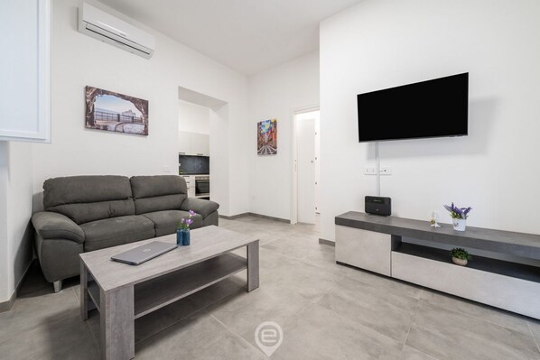 A modern living room with a grey sofa, a wooden coffee table, a flat-screen TV mounted on the wall, and a kitchen area in the background.