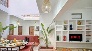 A modern living room with a dining area, featuring a TV, shelves with decorative items, and a large plant. - Riad Louzaia Marrakech (Marrakech)