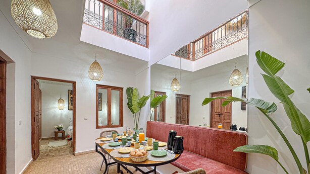 A dining area with a brick floor, a round table set with plates, cups, and a basket of bread, and a red bench. - Riad Louzaia Marrakech (Marrakech)