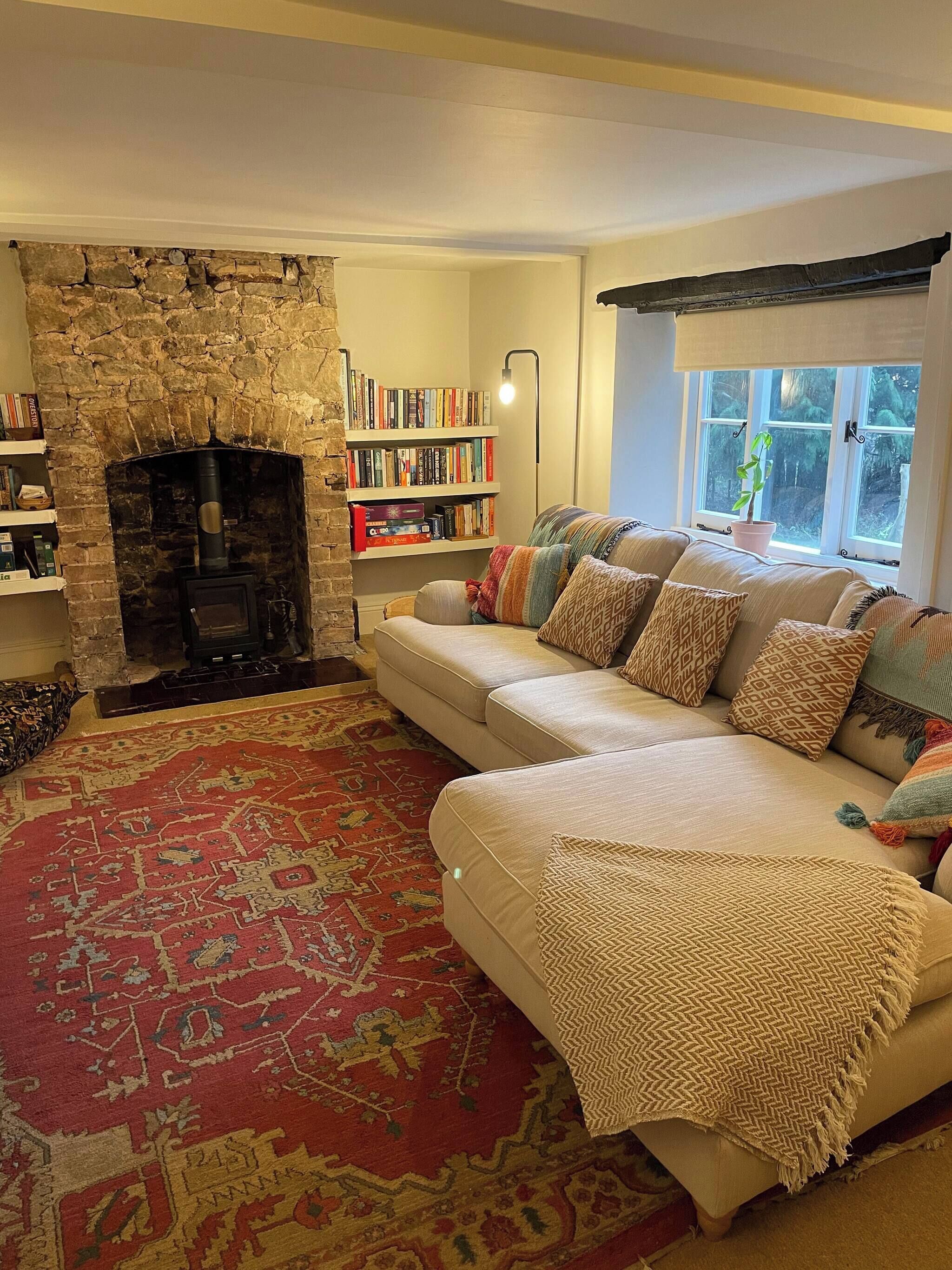 A cozy living room with a stone fireplace, a beige sofa with patterned cushions, a red patterned rug, and a bookshelf filled with books.