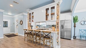 A kitchen with white cabinets, a central island with bar stools, a stainless steel refrigerator, and a hanging plant. - Good Vibes: New Mgmt 2026, Relaxing Private Escape w/ Pool & Beach Service (Destin)