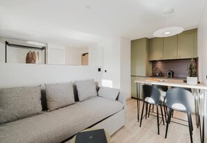 A modern living room with a sofa, a dining area with chairs, a kitchen with a counter, and a hanging light fixture. - Neostelia (Annecy)