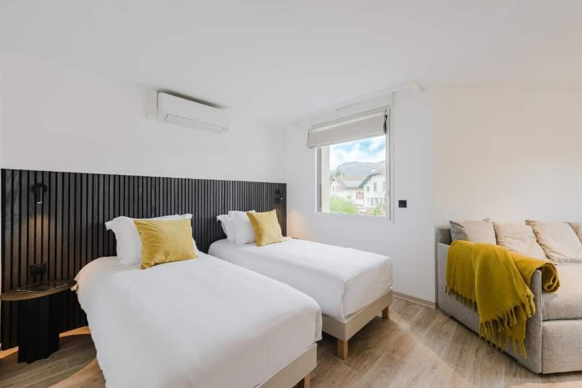Hotellihuone, jossa on kaksi sänkyä, sohva, ikkuna, josta on näkymä, ja seinälle asennettu ilmastointilaite.