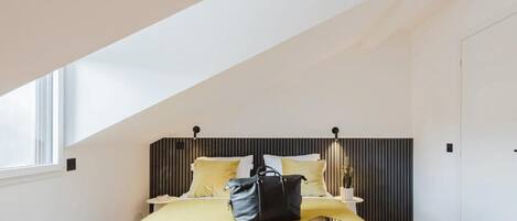 A bedroom with a sloped ceiling, a bed with a yellow bedspread, a black bag on the bed, and a bedside table with a lamp.