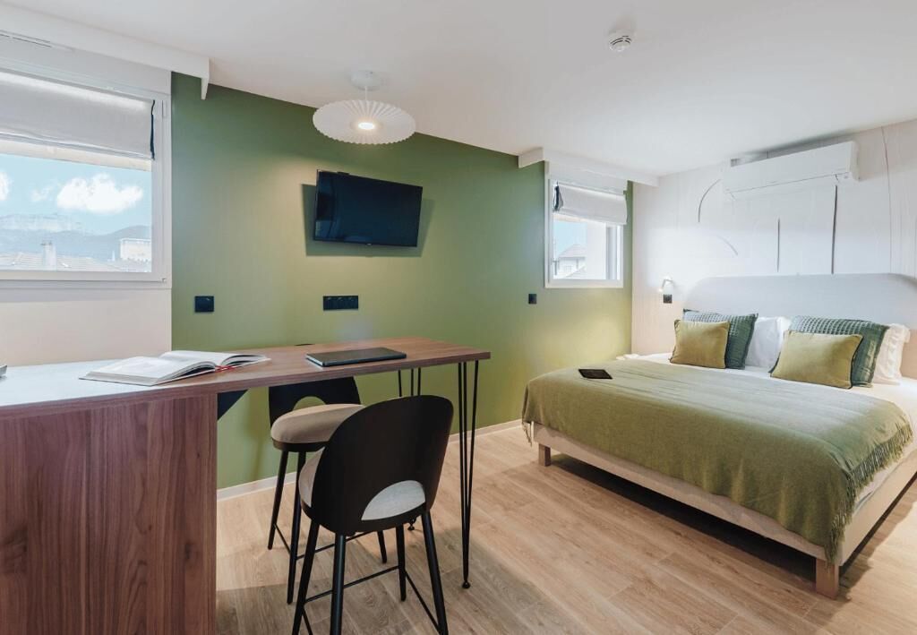 A bedroom with a bed, a desk, a chair, a television, and a window.