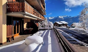 A snow-covered street with a wooden building featuring balconies and a restaurant. - Charming Vintage Flat Near Spa & Slopes (Leytron)