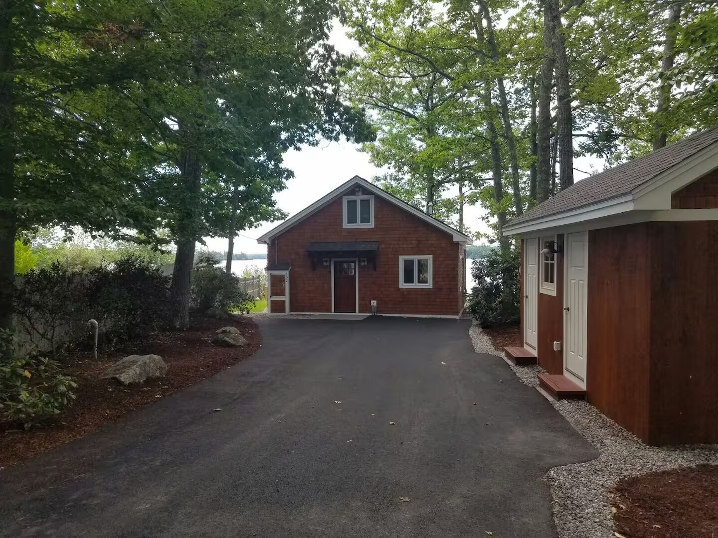 A two-story wooden house with a garage, surrounded by trees and a gravel driveway.