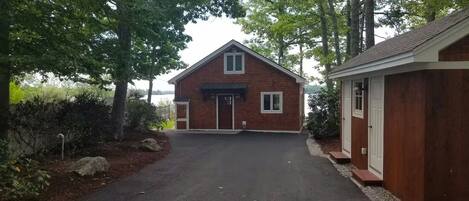A two-story wooden house with a garage, surrounded by trees and a gravel driveway.