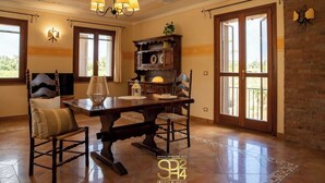 Un comedor con mesa de madera, sillas y una araña. Se ve una ventana con cortinas y vistas a la vegetación.