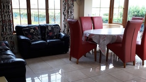 A living room with a sofa, a dining table with chairs, a TV mounted on the wall, and large windows with curtains. - Ballyferriter Dingle Seaside Beautiful House (Ballyferriter)