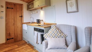 A compact kitchen with wooden cabinets, a sink, and a coffee maker. There is a chair with a checkered pillow and a framed picture on the wall. - Daves Place (Much Wenlock)