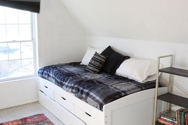 A bedroom with a white bed, a window, a bookshelf, and a patterned rug.