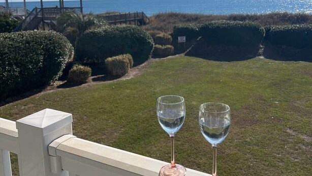 A balcony with two empty wine glasses, a view of the ocean, and a wooden gazebo in the background. - Isle Be Back - Oceanfront Ocean Isle (Ocean Isle Beach)