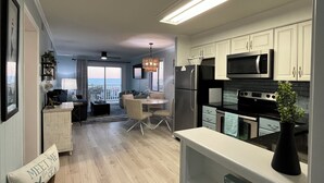 A modern kitchen with white cabinets, a black refrigerator, and a microwave. There is a dining area with a table and chairs, and a view of the ocean through large windows. - Isle Be Back - Oceanfront Ocean Isle (Ocean Isle Beach)