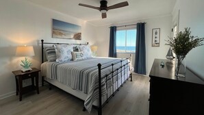 A bedroom with a bed, bedside tables, a nightstand, a ceiling fan, a window with curtains, and a view of the beach. - Isle Be Back - Oceanfront Ocean Isle (Ocean Isle Beach)