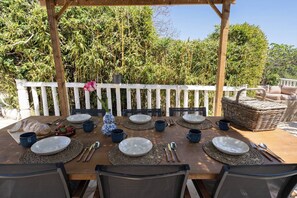 A wooden table set for a meal with plates, cups, and cutlery, surrounded by chairs, under a wooden pergola with a white fence in the background. - Casa Rosa Longterm Living and Workspace (Torrent)