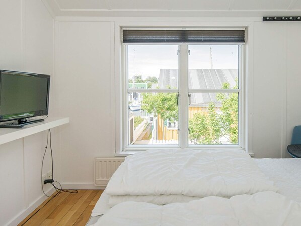 A bedroom with a bed, a television on a shelf, and a window with a view of buildings and trees. - 7 Person Holiday Home in Ebeltoft (Ebeltoft)