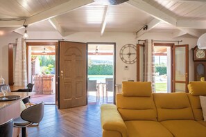 A room with a wooden ceiling, a yellow sofa, a dining table with chairs, and a view of an outdoor area with a pool. - Villa Serena - Alghero (Alghero)