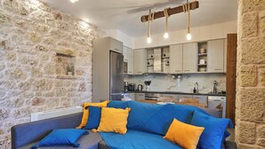 A modern living room with a stone wall, a grey sofa with blue and yellow cushions, and a kitchen area with white cabinets and a marble countertop. - Porto Santo Mani 1st Apartment (Mani)