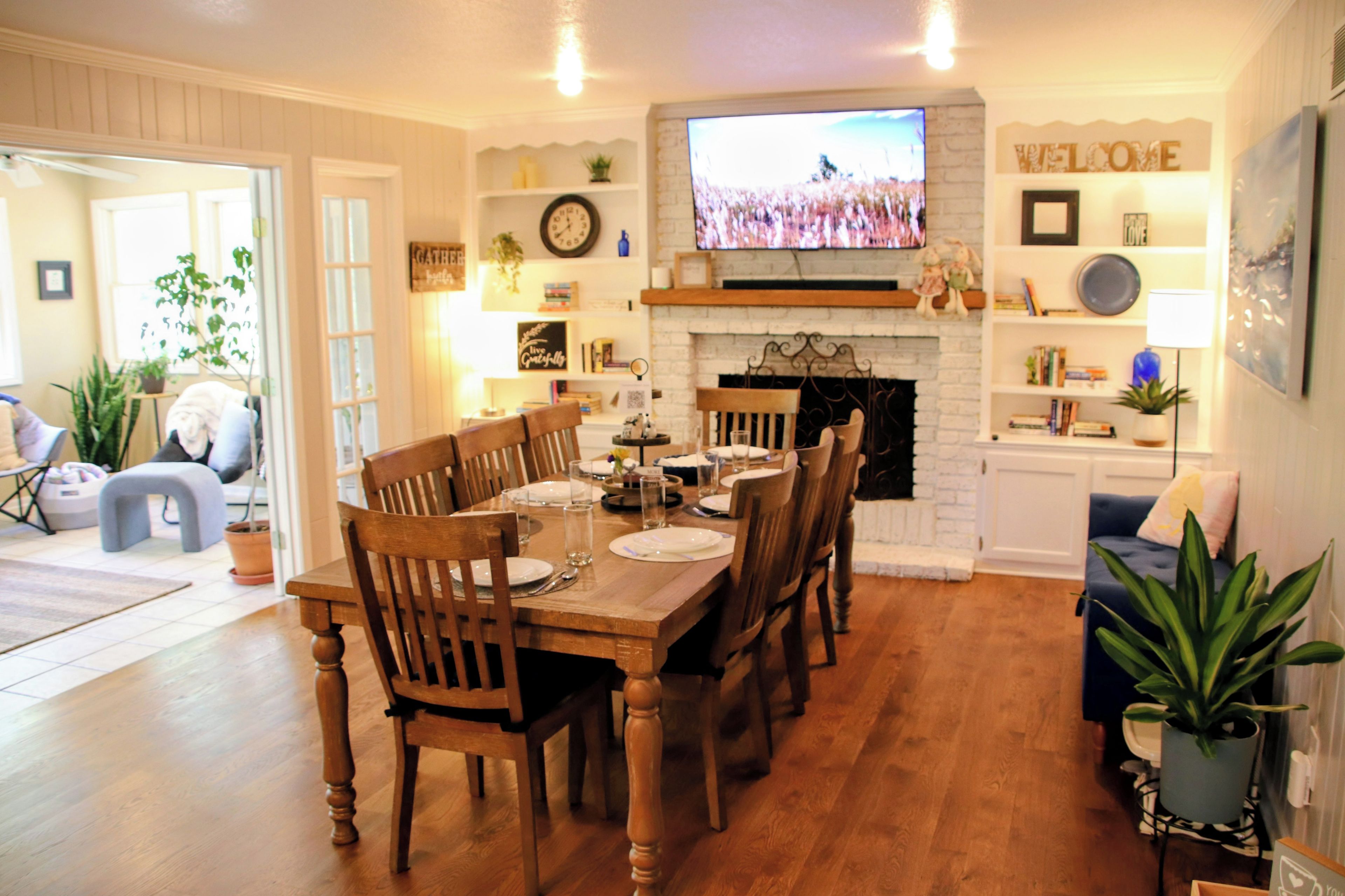 A dining area with a wooden table set for six, flanked by chairs, a fireplace, and a television mounted above.