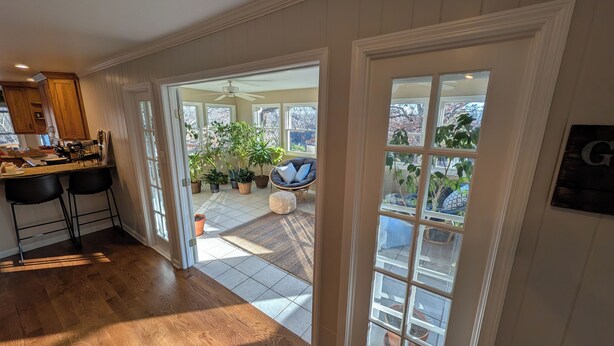 A kitchen with wooden cabinets, a countertop, and a dining area with two black chairs. There is a large window with a view of the outside, including a patio with potted plants and a chair.