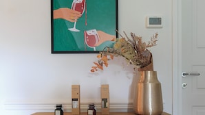 A wooden cabinet with a framed artwork of hands holding wine glasses, a vase with dried flowers, and two small containers on top. - Sense Rooms - Joy (Baveno)