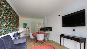 A modern living room with a grey sofa, a red rug, a dining area with a round table and chairs, a kitchen area with a black countertop, and a TV mounted on the wall. - Sense Rooms - Joy (Baveno)