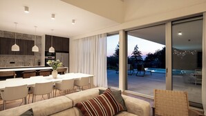 Una cocina moderna con iluminación colgante, un comedor con una mesa y sillas blancas, y un gran ventanal corredero que da a un patio exterior con piscina.