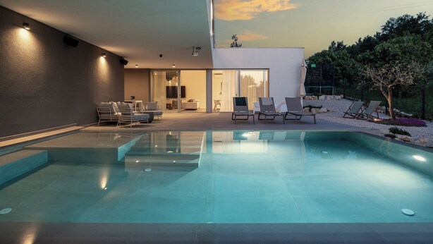 Un área de piscina moderna al aire libre con sillones de descanso y un espacio interior bien iluminado.