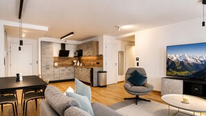 Ein modernes Wohnzimmer mit grauem Sofa, einem Esstisch mit Stühlen, einer Küchenzeile mit Holzschränken und einem großen Fernseher an der Wand.