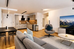 A modern living room with a grey sofa, a dining table with chairs, a kitchen area with wooden cabinets, and a large TV on the wall. - Apartment Aiguille d'Or (Les Houches)