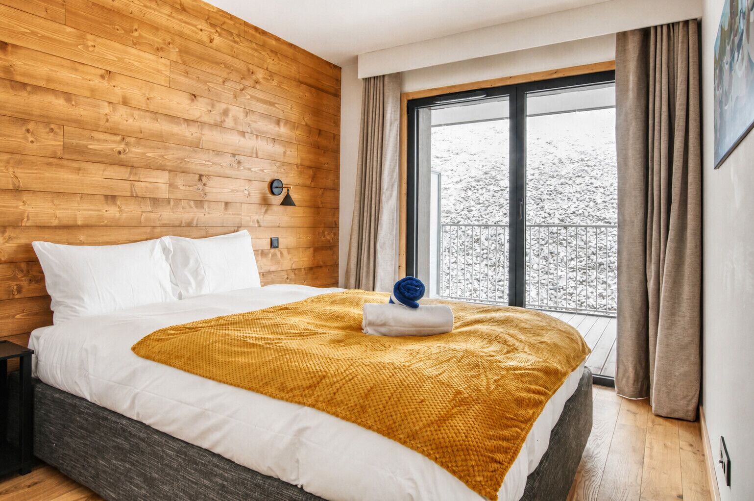 A hotel room with a wooden wall, a large bed with white bedding, a yellow throw blanket, and a view of a snowy landscape through a window.