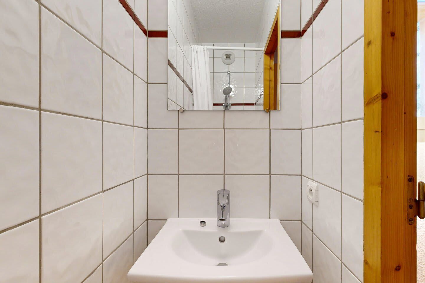 Une salle de bain avec des murs recouverts de carreaux blancs, un lavabo blanc et un miroir.