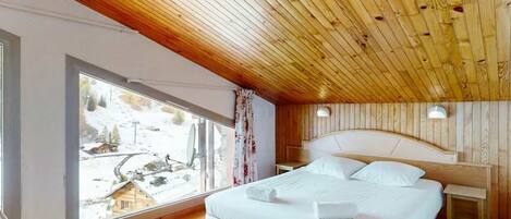 Un dormitorio con techo de madera, un ventanal con vistas a un paisaje nevado, una cama con ropa de cama blanca y una mesita de noche con una lámpara.