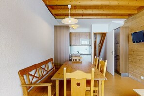 Une table à manger en bois avec des bancs, un coin cuisine équipé d’un four à micro-ondes et un téléviseur fixé au mur.