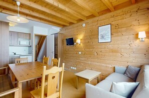 L’intérieur chaleureux d’un chalet avec une table à manger en bois, des chaises, un canapé et un téléviseur fixé au mur.