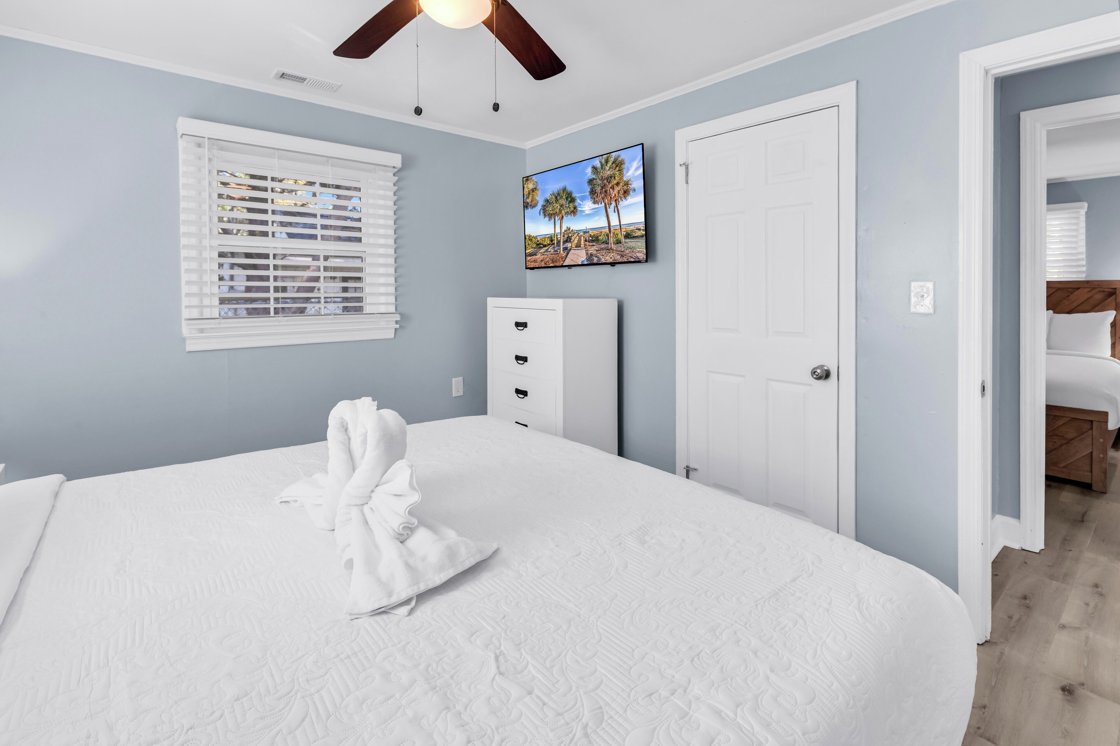 Une chambre avec un grand lit, un ventilateur de plafond, une fenêtre avec des stores, une porte et un téléviseur fixé au mur.