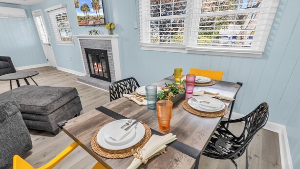 A dining area with a wooden table set for four, surrounded by modern chairs, a fireplace, and a view of palm trees through the windows. - Sea Breeze Retreat (North Myrtle Beach)