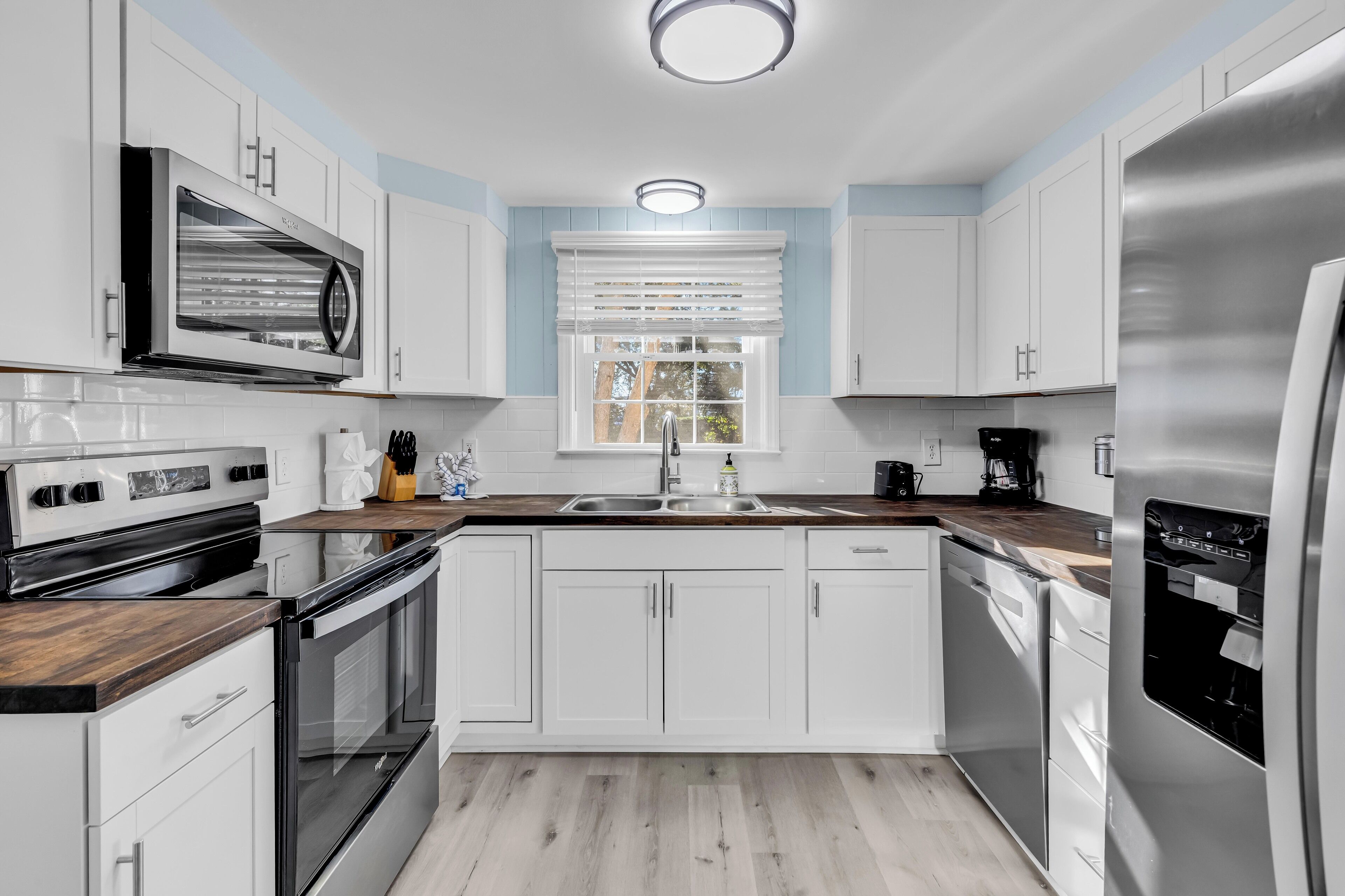 Une cuisine moderne avec des meubles blancs, des appareils en acier inoxydable, un plan de travail en bois et une fenêtre équipée de stores.